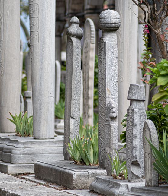 A cemetery scene in Nunhead Cemetery illuminated by soft morning sunlight, showing a variety of weathered headstones and memorial statues made of stone and marble, some with intricate carvings and ins